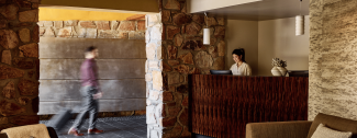 Hotel lobby with stone walls, a man walking with luggage, and two people behind a wooden reception desk.